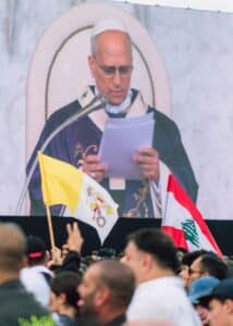 Man in religious attire reads from papers on screen.