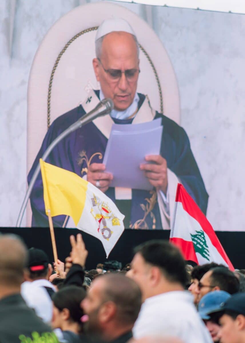 Man in religious attire reads from papers on screen.